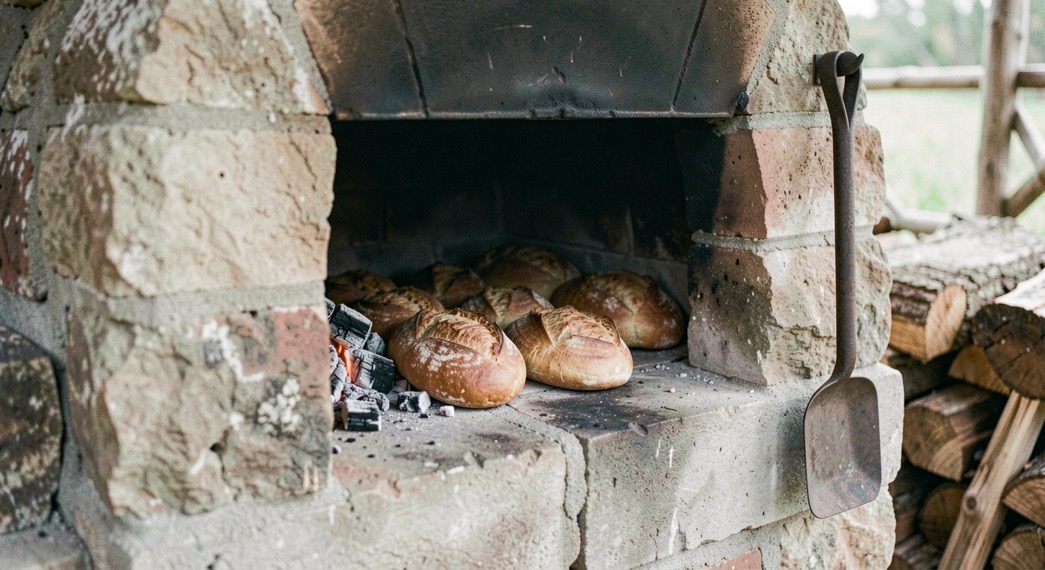 Boca de horno de leña de obra abierta con las brasas arrimadas a un lado, panes ya dorados dentro sobre la piedra y un atizador de hierro colgado al lado